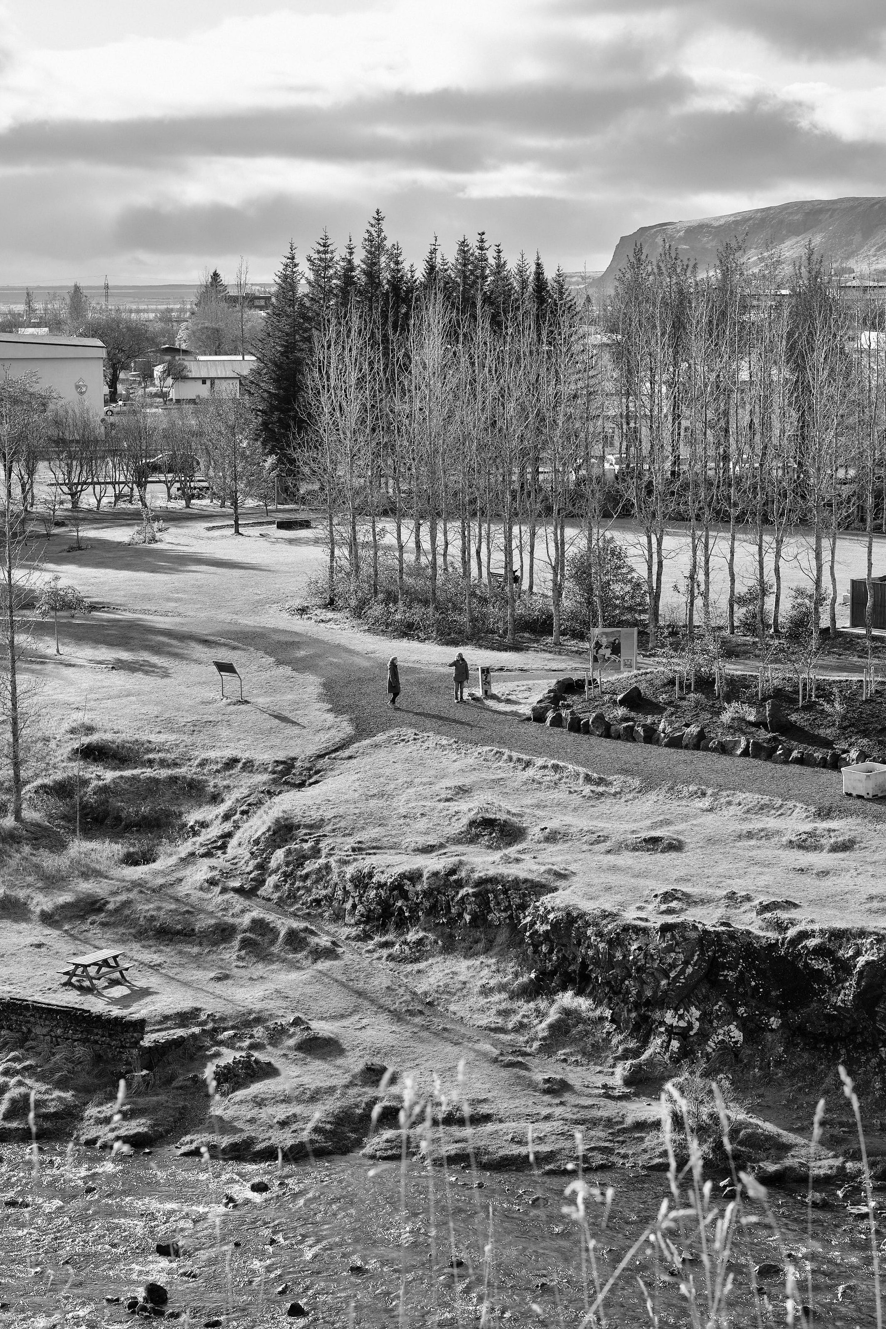 In the distance, two people are walking together through a park in Hveragerði. One is pointing something out for the other.